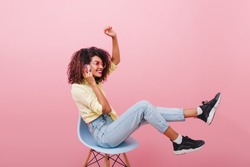 Glad brunetteIndoor full-length portrait of confident african girl in pink shirt holding skateboard. Enthusiastic black woman with curly hairstyle posing in studio with blue interior. in black sneaker