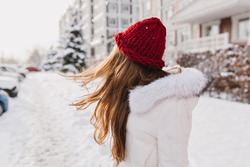 Portrait from back of amazing girl posing with long hair waving in cold winter day. Outdoor photo of dreamy woman in woolen red hat looking in distance standing at snowy street.