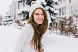 Close-up portrait of happy girl in woolen sweater enjoying winter moments. Outdoor photo of long-haired laughing lady in knitted hat having fun in snowy morning on blur nature background.