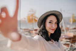 Dreamy brunette girl with short hair expressing happy emotions while making selfie outdoor. Photo of elegant young woman in hat and blouse taking picture of herself on blur background.