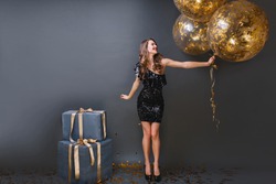 Full-length portrait of refined european girl wears black dress at birthday party. Blissful long-haired lady with balloons can't wait to open presents.