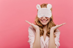 Studio shot of pleased beautiful young woman posing in eyemask. Cheerful european girl in pajamas standing on pink background in sleep mask in morning.