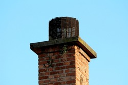 Dilapidated old red building brick chimney with plants growing from cracks and concrete top covered with dense rusted wire mesh cover on clear blue sky background