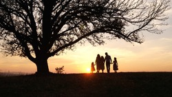 Silhouettes of happy family walking in the meadow near a big tree during sunset. Family spent time together. Enjoing each other