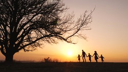 Silhouettes of happy family holding the hands and walking together in the meadow near a big tree during sunset