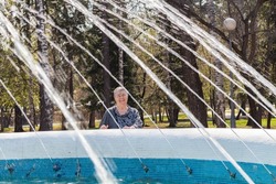 senior woman sincerely smiles looks at camera standing by fountain. Water jets and water splashes frame pensioner's face outdoors. sun rays illuminate attractive woman and mosaic fountain.