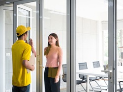 Asian delivery man who wear yellow uniform with cap hold confidential document in craft paper and knock the window for send envelope to customer in meeting room at corporate office