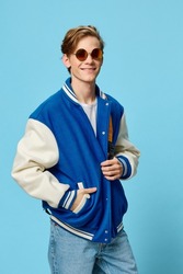 a happy, handsome, fashionable student in a blue bomber jacket is standing in sunglasses and with a bright backpack on his back. Vertical studio photography