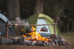 Camp fire and tea pot are foreground and focused, there is a tent in the background and defocused