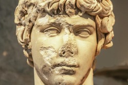 Close-up of head of ancient marble statue of attractive Greek youth with parts of face chipped off and destroyed