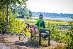 Long haired person in blue jeans with backpack and helment sits looking out at bay from park bench near downtown Anchorage Alaska in summertime