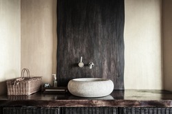 Modern bathroom interior with original white stone sink, clock, wooden baskets and soap dispenser. Wooden interior of spa in yellow and gold colors. Selective focus, only part of bathroom is in focus.