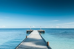 Perspective view of a wooden pier on the tropical seashore with clear blue sky with some white clouds and sea with turquoise water.