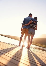 The end to a perfect day together. Rearview shot of an affectionate young couple standing on a dock at sunset.