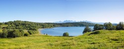 Landscape of a lake with trees near a field. Green hills by the seaside with a blue sky in Norway. A calm lagoon near a vibrant wilderness against a bright cloudy horizon. Peaceful wild nature scene