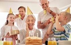 Senior woman celebrating her birthday with family at home, wearing party hats and blowing whistles. Grandma looking at birthday cake and looking joyful while surrounded by her grandkids and and son
