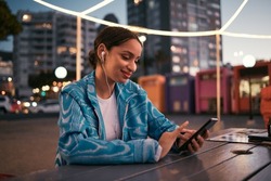 Happy and trendy woman browsing on phone while wearing earphones and listening to music while sitting at outdoor cafe in night city. Happy woman streaming subscription app or making video call