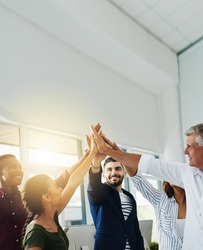 Group of business people doing high five together while standing inside an office with sun flare and copy space. Team of colleagues, coworkers and employees celebrating teamwork and stacking hands