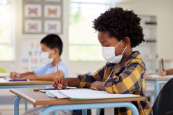 School student in class during covid pandemic for learning, education and study with mask for safety, protection and protocol. Little kindergarten, preschool or elementary kid writing in book at