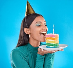 Celebrate, birthday and woman in studio with a cake, party hat and candle for a celebration. Happy, smile and Indian female model ready to eat sweet rainbow dessert while isolated by blue background.