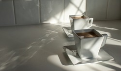 Square white tea cups against a white tiled wall. Good contrast between black and white. Square cups and tiles in shadow and light make a unique kitchen image with strong contrast of shape and light