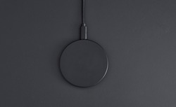 High angle view of black plastic circular wireless charger laying on dark grey desk. Modern technology, wireless device and transfer of energy concept.