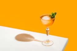 Elegant stemware glass of fresh orange cocktail with big ice ball on white table surface, bright yellow background. Spring summer art drink food concept.