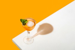 Elegant stemware glass of fresh orange cocktail with big ice ball on white table surface, bright yellow background. Spring summer art drink food concept.