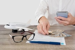 Man with smartphone applying travel insurance form with plane model, glasses and cash money lying on table, studio, light gray background