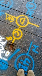Alphanumeric and symbol spray paint markings on a pedestrian footpath to indicate different types of construction works needed. Pair of booths showing in the lower image.