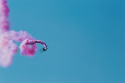 Aerobatic plane leaving a pink smoke trail in the bright blue sky.