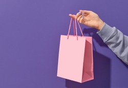 Womans hand holding pink paper shopping bag on purple background. Shopping sale delivery concept