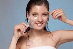 young and happy female peeling off facial mask and smiling to the camera, woman treats her face in bath towel
