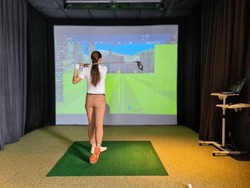 Professional female golfer holding club playing golf indoors on golf simulator. Driving range with virtual golf screen