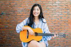 Beautiful Asian female musician performing live in front of audience recording viral video on social media singing playing guitar taking to people, camera vlogging at working at home brick background 