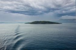 the wake of a sea bound cruise ship in the distance a tree covered island under a brooding Mediterranean sky