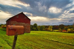 Ghent, NY, USA - Sep 27th 2020: red mailbox on side of road. A red mailbox stands on the side of the road with a red barn and fields in the background. Trees are starting to change color.