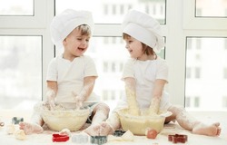 Happy children - a boy and a girl play in the kitchen with flour, children knead the dough, a child in a cook's costume, cooking from dough, playing cook.
