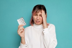 a woman suffering from a headache holds painkillers while standing on a blue background