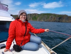 A middle-aged woman in glasses, a hat, a red jacket and blue jeans sits on board a boat and smiles. Leisure, recreation, travel, tourism.
