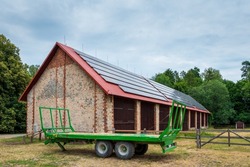 Green farm trailer parked in front of a large barn. Solar panels installed on the roof of the barn