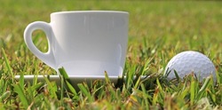 White cup of coffee next to a golf ball, on a green lawn