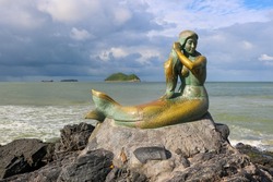 A mermaid statue sitting on a rock by the sea​ with a small island in the background