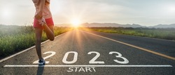 New year 2023 or start straight concept.word 2023 written on the asphalt road and athlete woman runner stretching leg preparing for new year at sunset.Concept of challenge or career path and change.