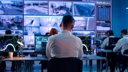 Back view of young man with headset watching surveillance footage and map on computers and speaking with female colleague while working in intelligence agency