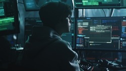 Young hacker drinking hot beverage and checking malicious code on computer monitors while sitting at desk in dark room at night
