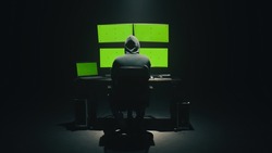 Concept shot of anonymous man in hoodie writing code to hack database while sitting at desk with computer monitors, with green screens chromakey in dim room of hacker base