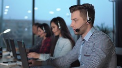 Young handsome men in checked shirt and headset working in call center in modern worldwide office he talking and looking on screen of desktop computer