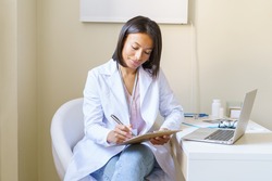 Young african woman doctor GP in white coat holding clipboard and filling patient form while working in medical clinic, afro american female medical worker doing paperwork at workplace with laptop