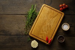 Food seasoning background. Spices, herbs and wood chopping board on dark wooden backdrop. Rosemary, thyme, salt, pepper, cherry tomato, garlic clove red chili. Menu, horizontal mockup, banner